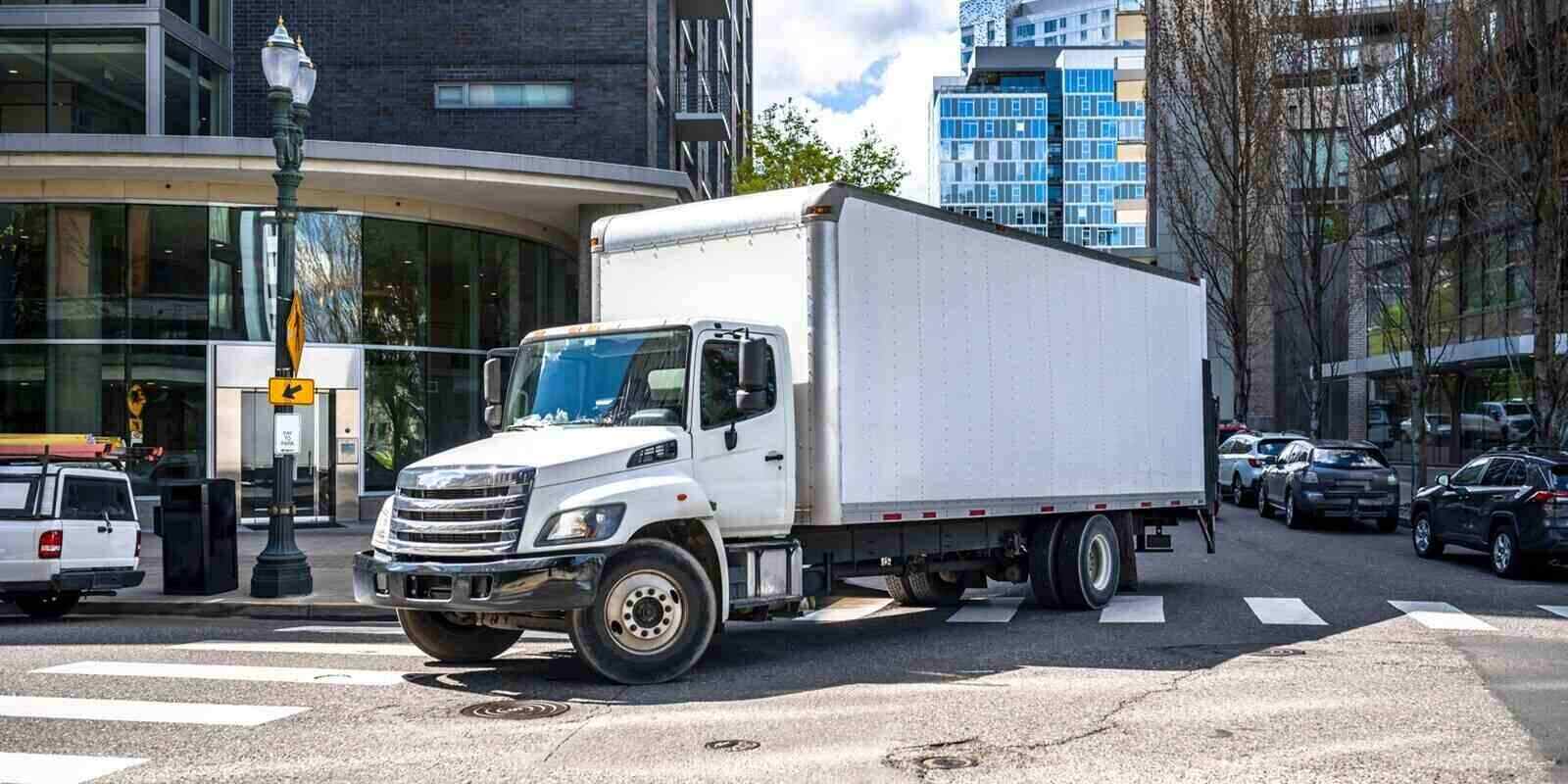 big rig day cab white semi truck with long box trailer making local commercial delivery at urban city with multilevel residential apartments buildings turning on the city street with crossroad
