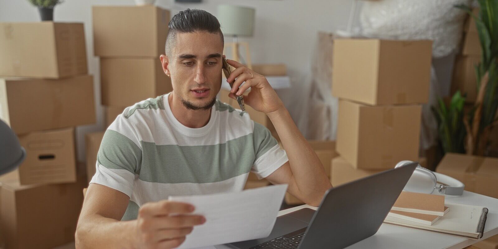 man holding paperwork and phone to ear while reviewing documents at a laptop in a new apartment filled with moving boxes
