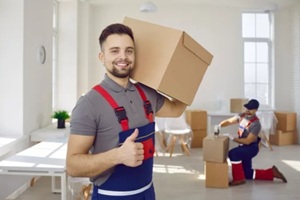 smiling male worker of moving and delivery company holding cardboard box showing thumbs up